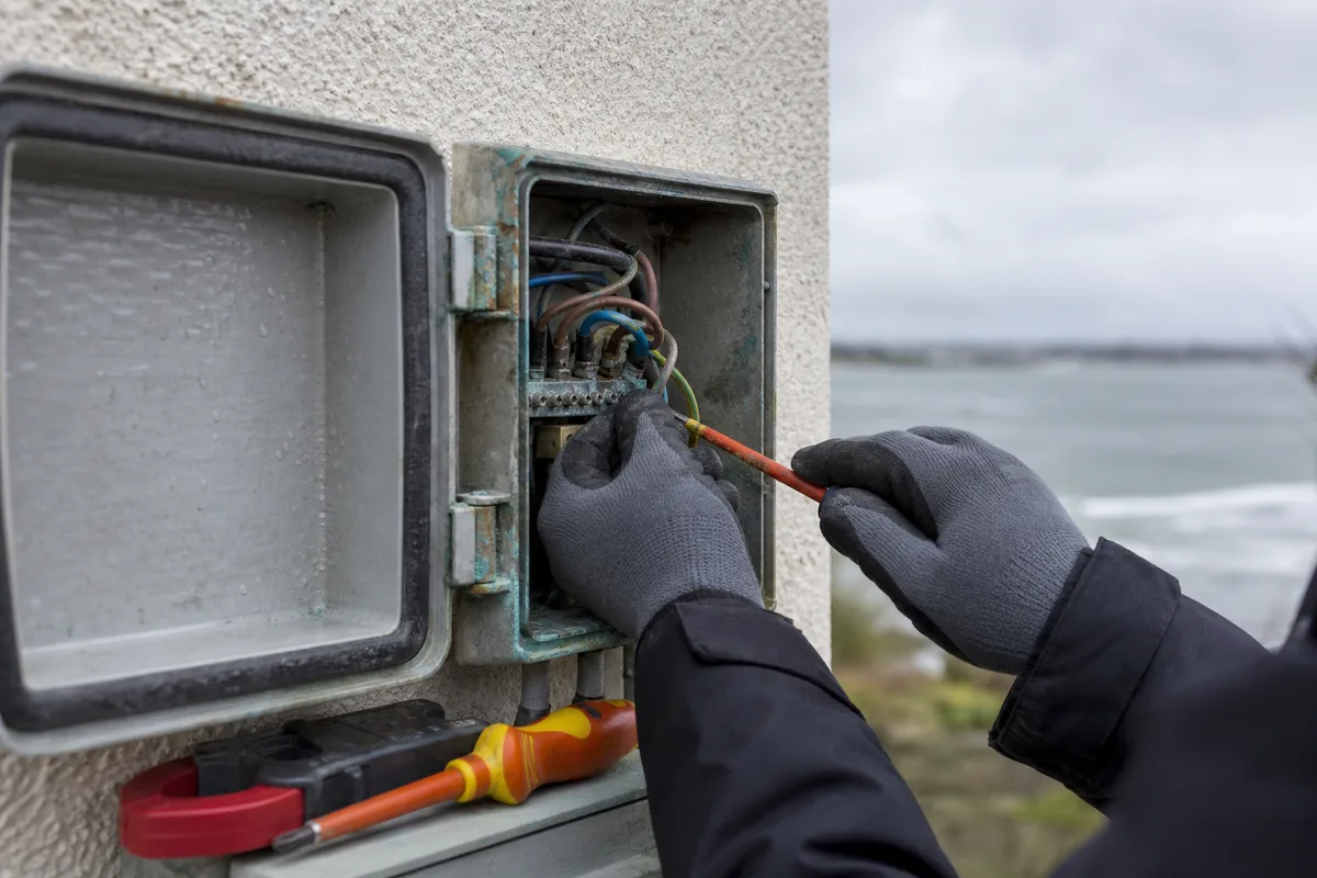 Mains d&rsquo;un électricien contrôlant les serrages d&rsquo;un coffret extérieur en bord de mer